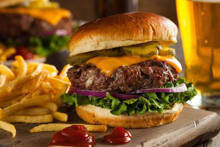 A juicy cheeseburger with lettuce, pickles, and purple onions, served with French fries and a beer.