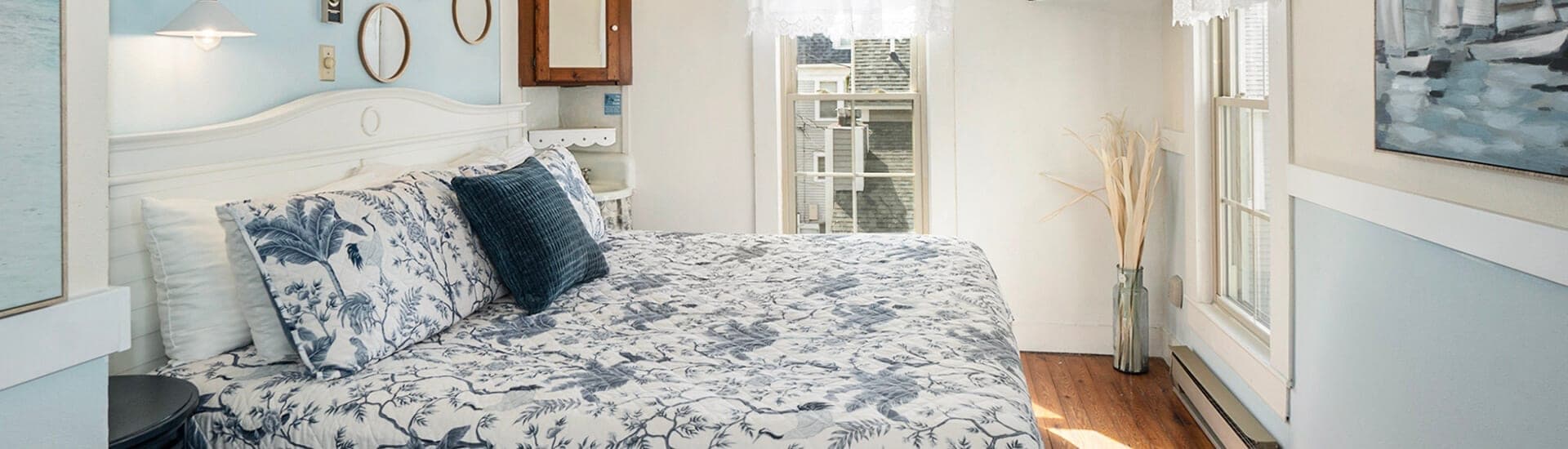A bright bedroom featuring a bed with blue and white patterned bedding, light walls, and natural light from windows.