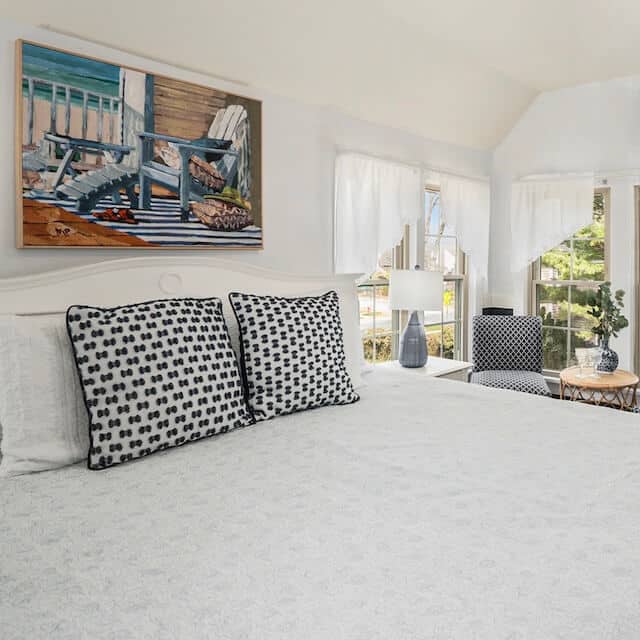 A cozy bedroom featuring a white bedspread, patterned pillows, and large windows with sheer curtains.