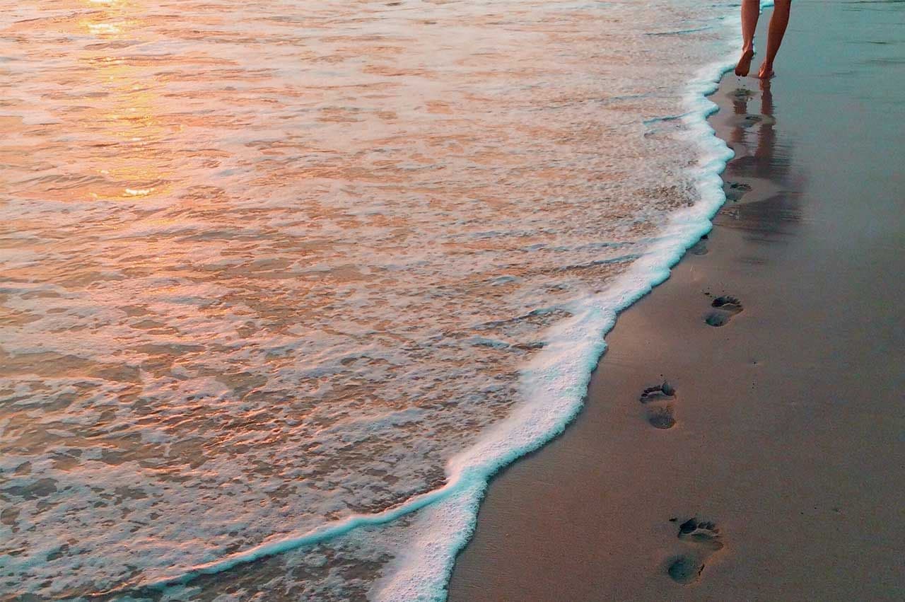 A person walks along a sandy beach with gentle waves lapping at their feet during sunset.