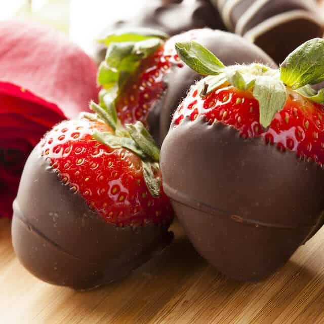 Chocolate-covered strawberries next to a rose on a wooden surface.