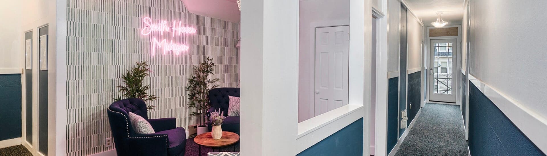A modern hallway leading to a cozy seating area with a neon sign that reads "Simply Home Mental."