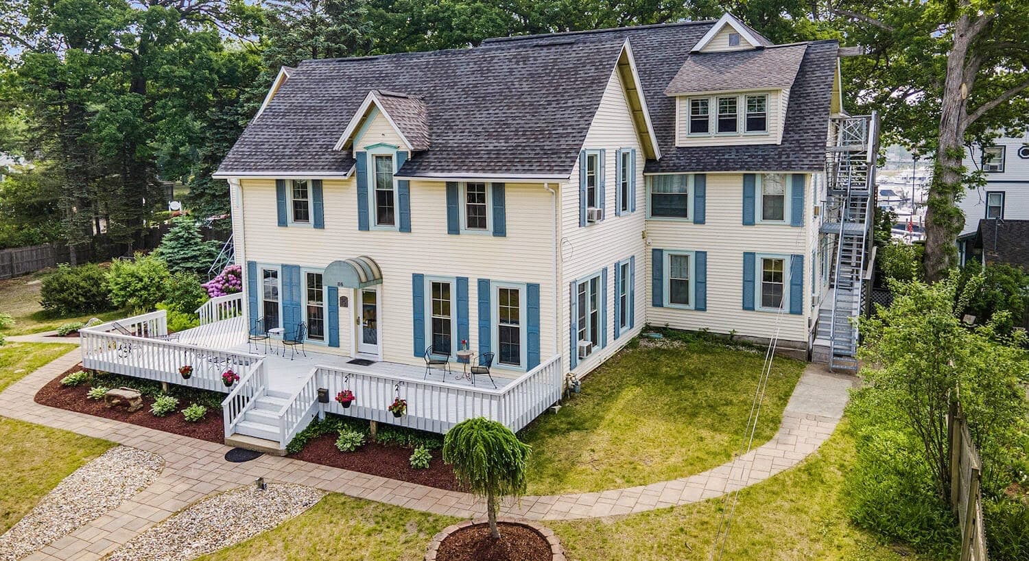 A large, two-story yellow house with blue shutters and a wraparound porch, surrounded by a landscaped yard.