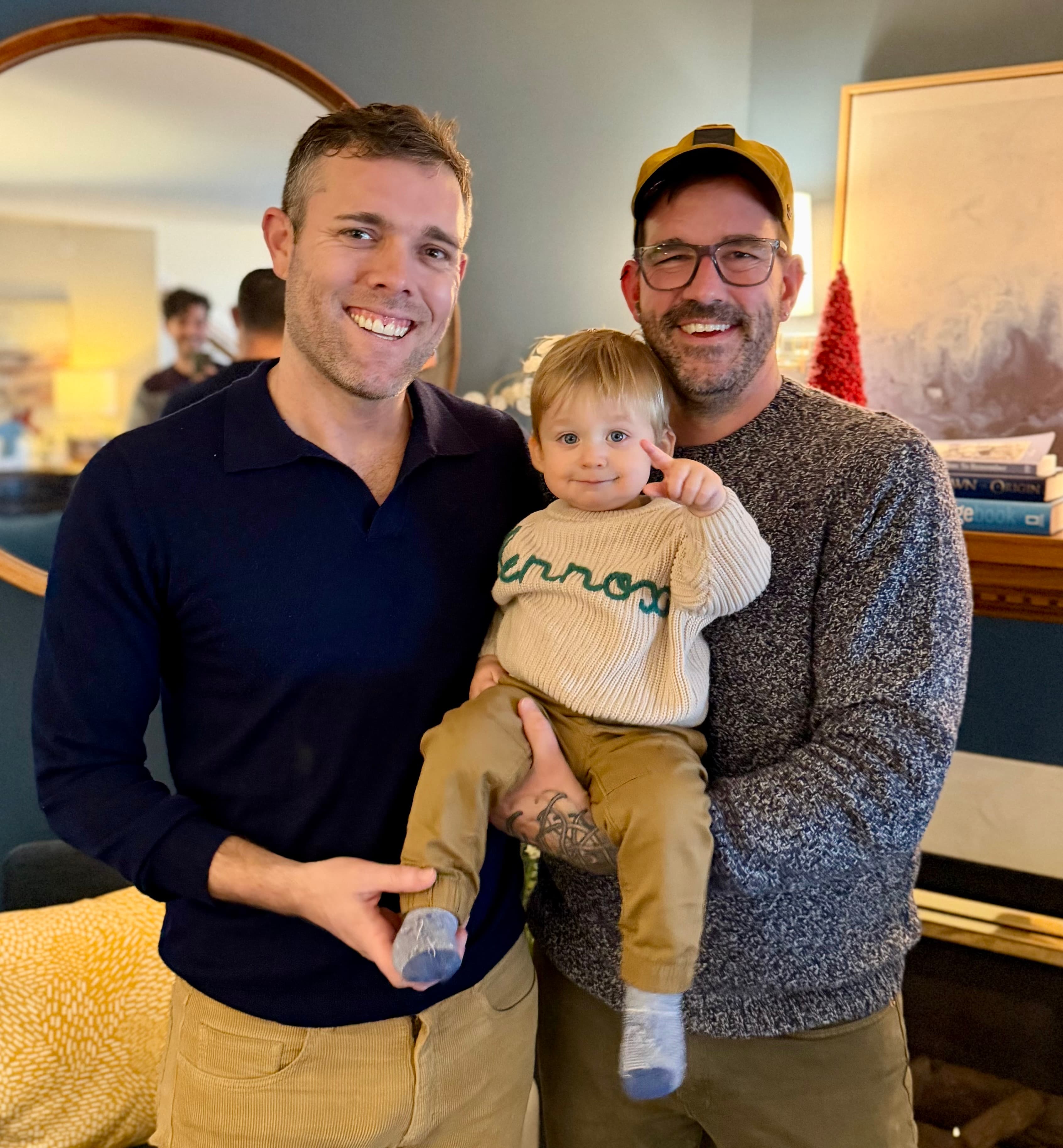 Two men stand together, one holding a toddler, with smiles in a cozy interior setting.