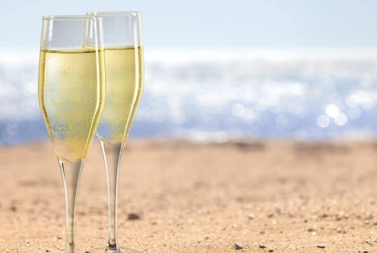 Two fluted glasses of sparkling champagne on a sandy beach with a blurred ocean backdrop.
