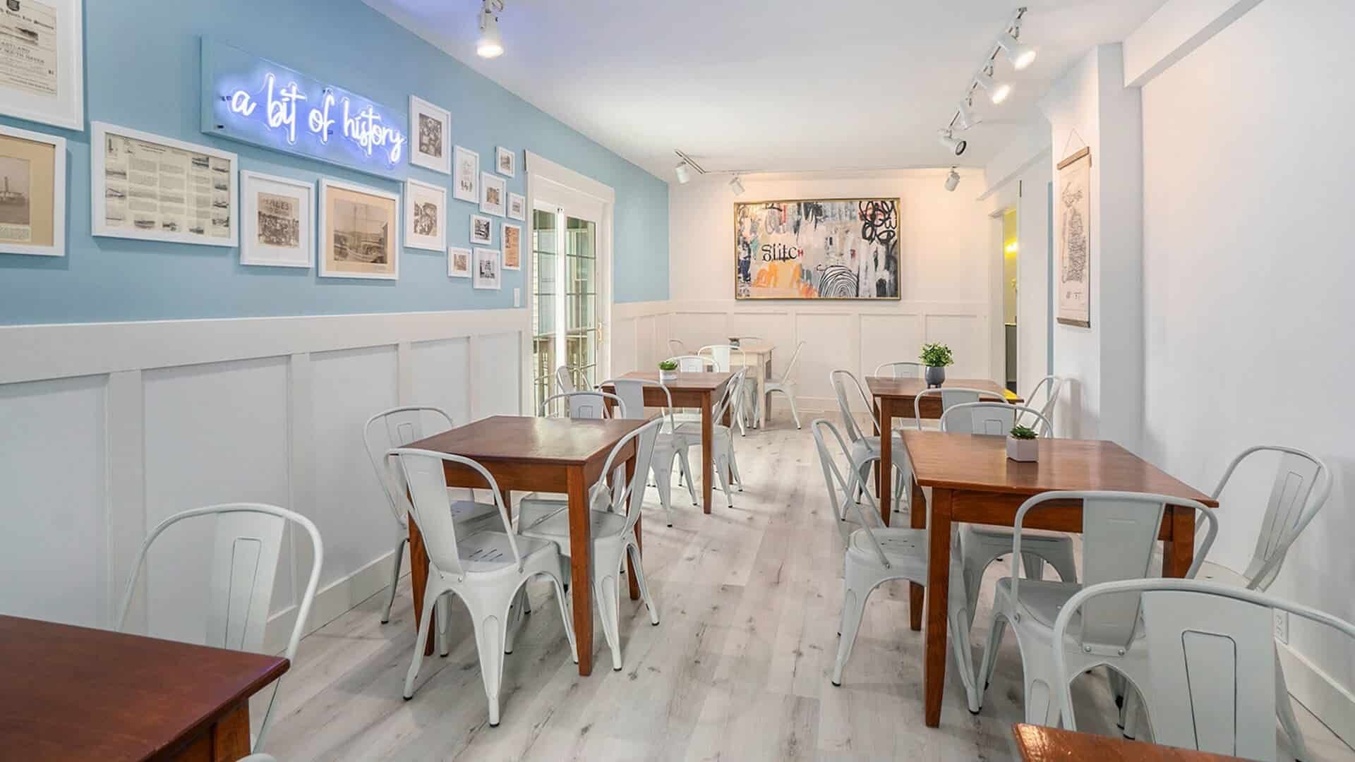 A bright café interior featuring wooden tables and metal chairs, with framed artworks on the walls and a neon sign.