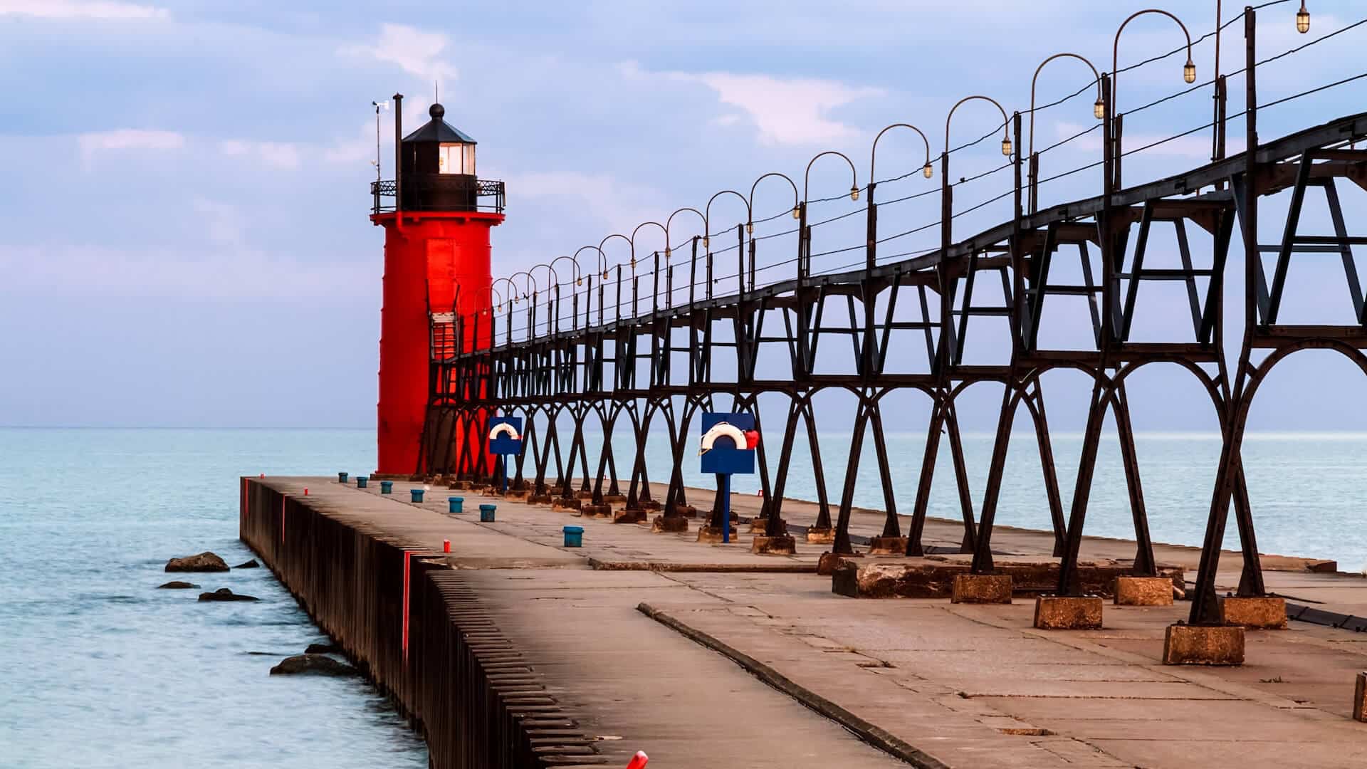 A red lighthouse stands at the end of a pier extending over tranquil water under a cloudy sky.