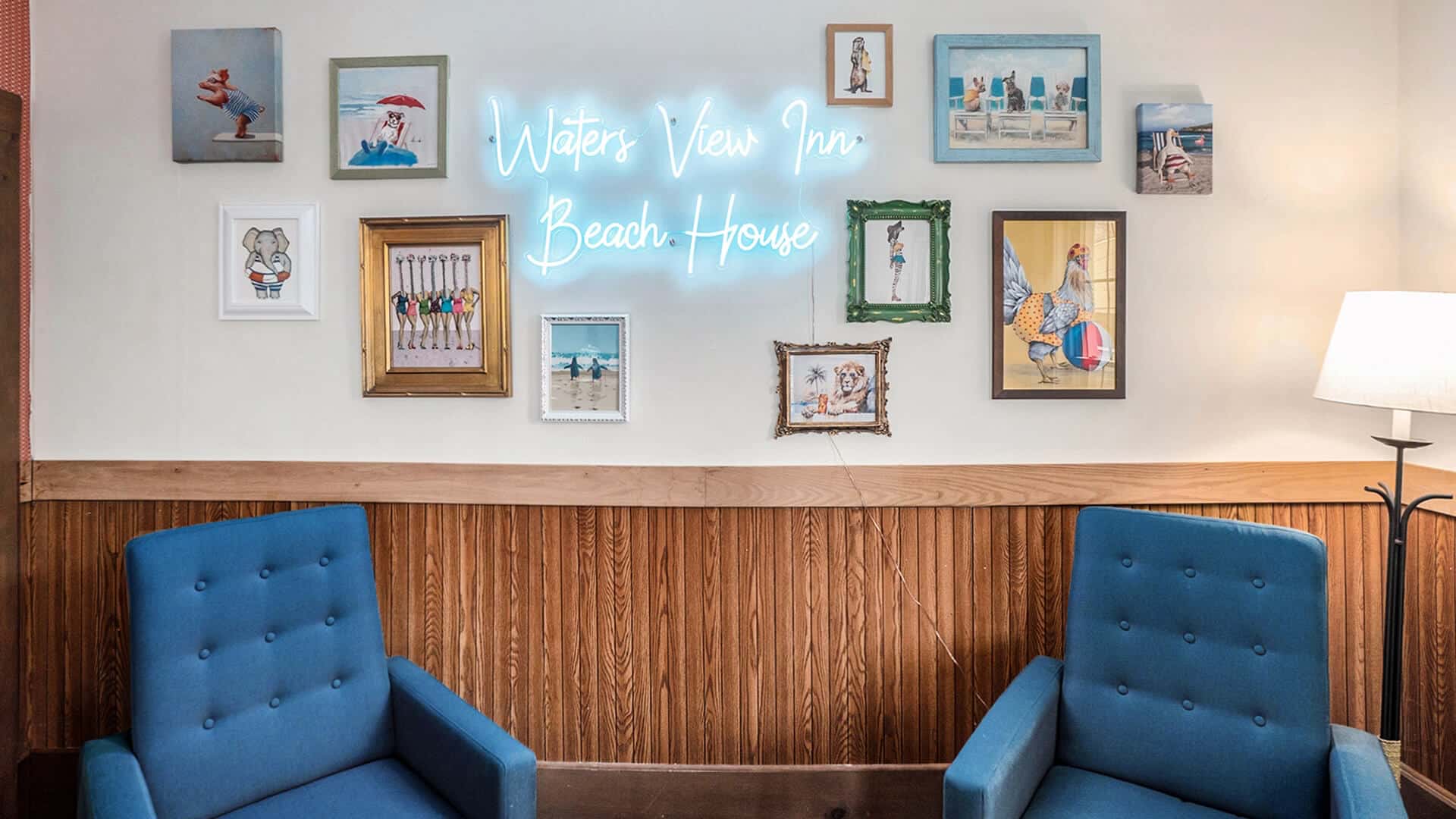 A cozy seating area with blue chairs and a gallery wall featuring beach-themed art and a neon sign that reads "Waters View Inn Beach House."