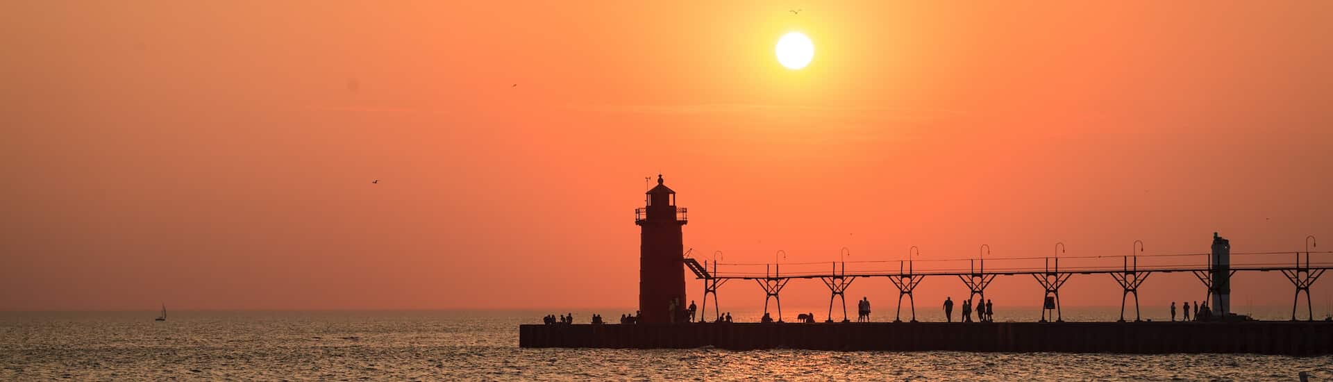 Sunset over a calm sea, with a silhouette of a lighthouse and people walking on a pier.