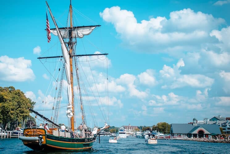 A tall ship sails through a harbor under a blue sky with fluffy clouds.