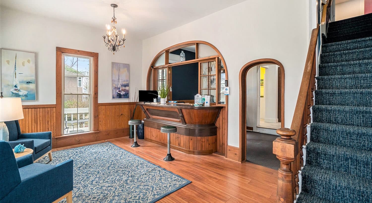 A sitting room with wood floors, white walls and wood wainscotting, an antique bar and barstools along one wall, and a sitting area with blue wingback chairs on the opposite wall, and stairs with blue carpeting leading upstairs.