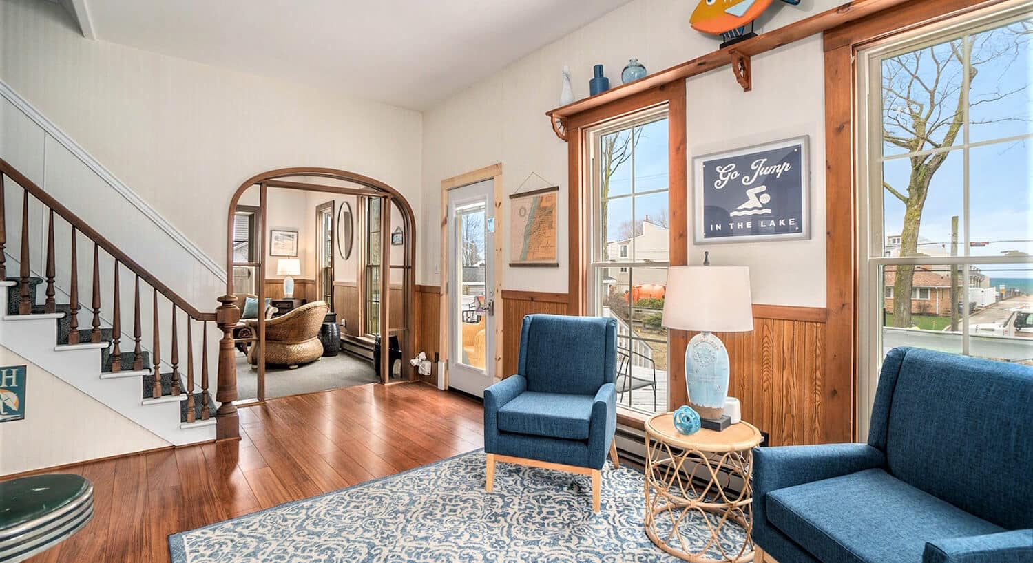 A parlor with wood floors, a blue and white tapestry area rug, blue wingback chairs, a wicker table with a lamp in between them, an archway leading to another sitting room, and stairs leading to a second floor.
