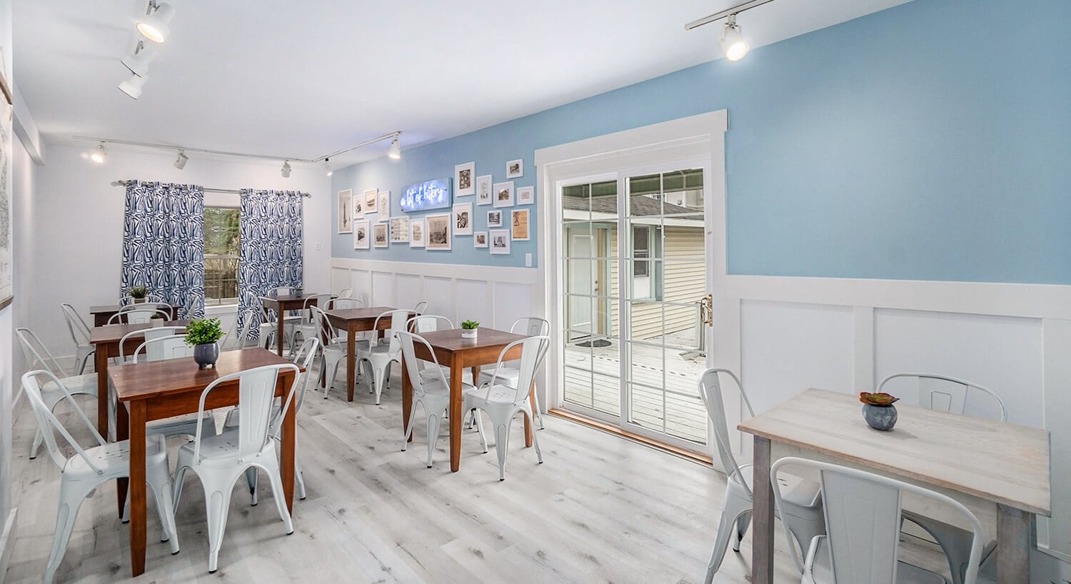 A dining room with pale blue walls, white wainscotting, wood floors, and several wood tables and metal chairs throughout the room.