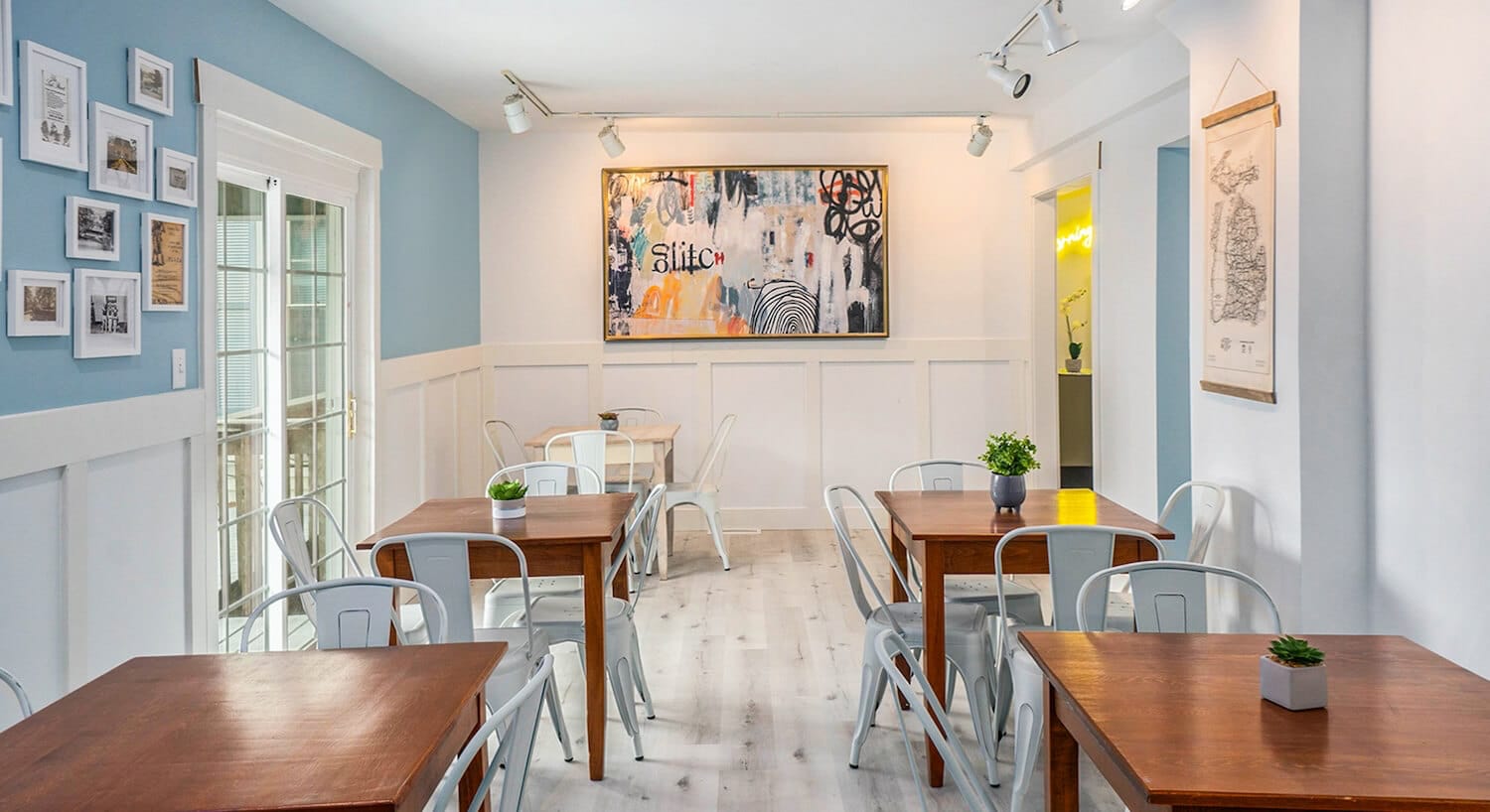 A dining room with pale blue walls, white wainscotting, wood floors, and several wood tables and metal chairs throughout the room.