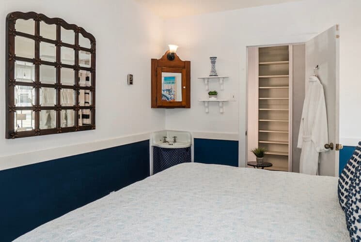 A bedroom with dark blue and white walls, a bed with white bedding, a sink in the corner, and an open closet with robes hanging on the door hooks.