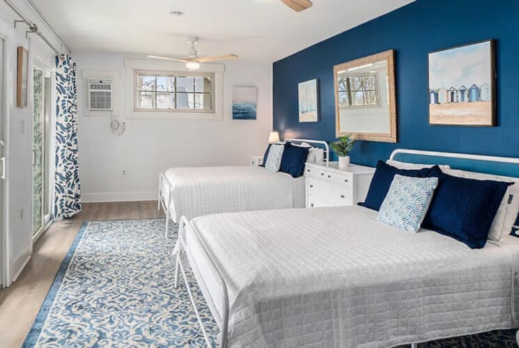 A bedroom with blue and white walls, wood floors, two large beds, a dresser in between the beds, nightstands on either end of the beds, and glass doors to an outside deck.