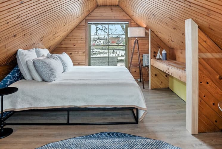 A top floor bedroom with knotty pine slanted ceilings, wood floors, a bed, tables with lamps, and a window along the front wall.