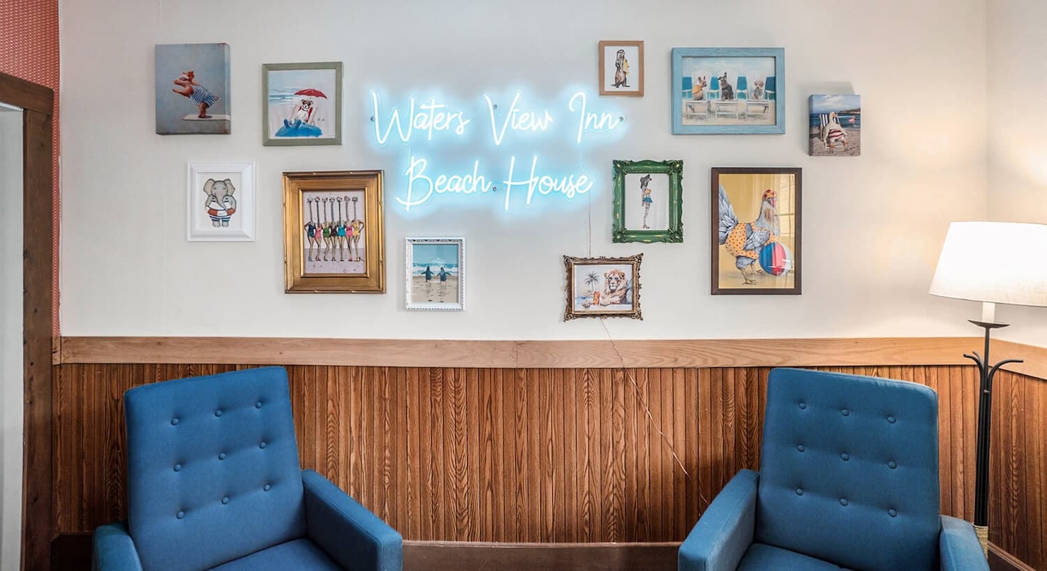 A sitting area with 2 blue armchairs along a wall with wood wainscotting, photos of animals on the wall, and a neon sign that says Waters View Inn Beach House.