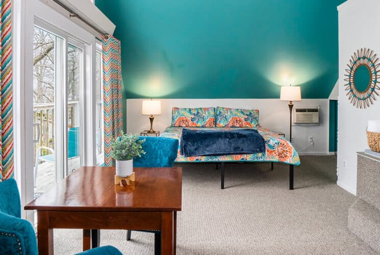 A bedroom with white and teal walls, a bed with colorful bedspread a wood table with two plush blue chairs, an adjacent built-in bathtub, and sliding doors leading to outside.