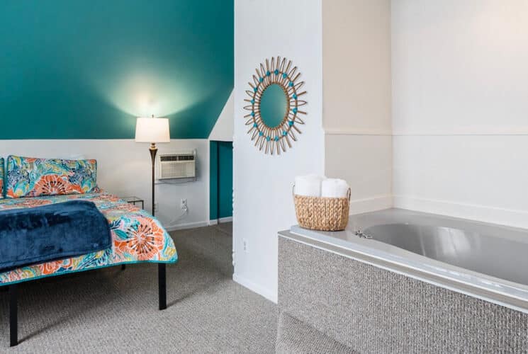 A bedroom with white and teal walls, a colorful bedspread and an adjacent built-in bathtub.