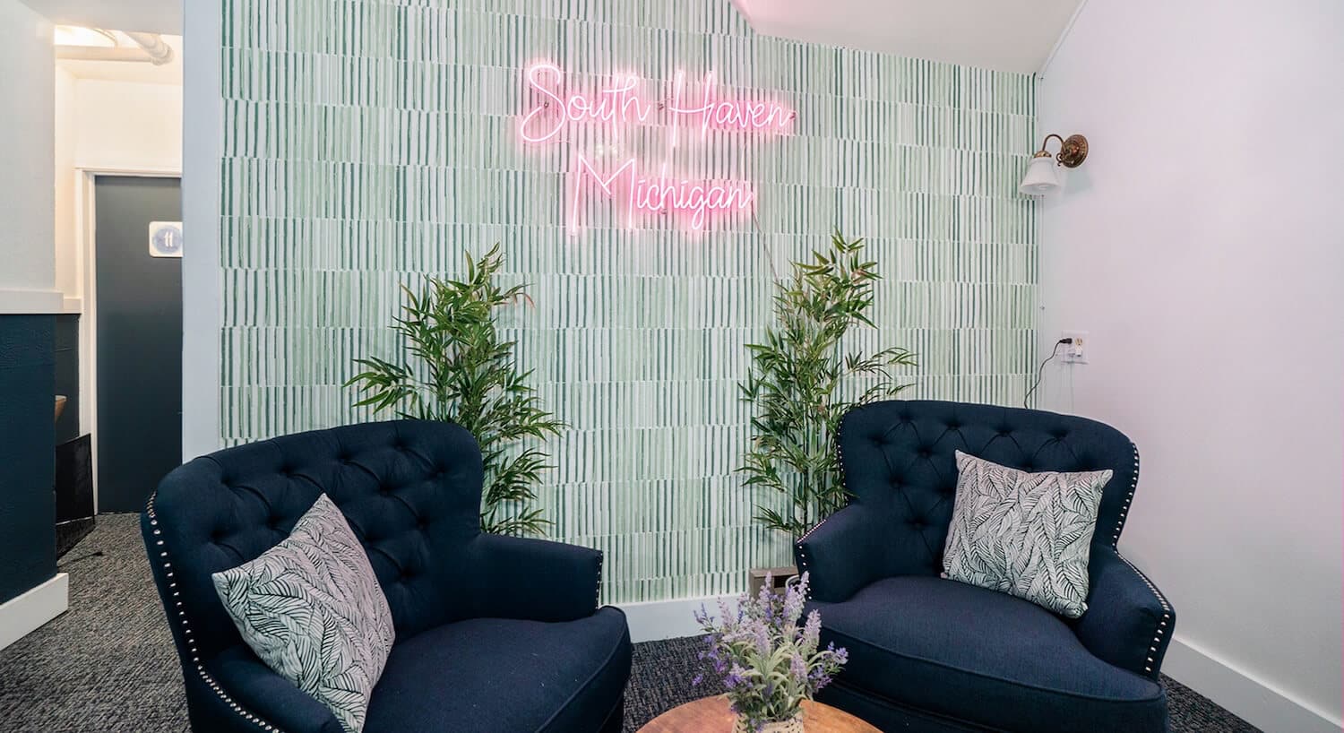 A sitting area in the corner of a room with plush dark blue wingback chairs, a wall with green wallpaper and a neon sign that says South Haven Michigan.