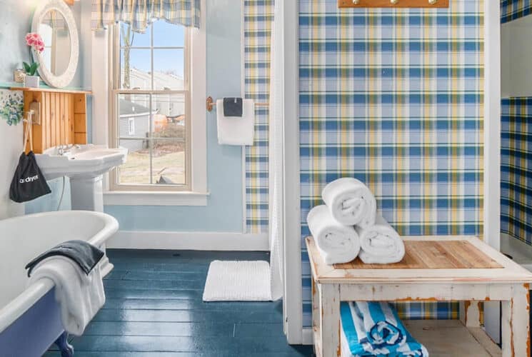 A bathroom with blue painted floors, a claw foot tub, with blue, yellow and white checked wallpaper.