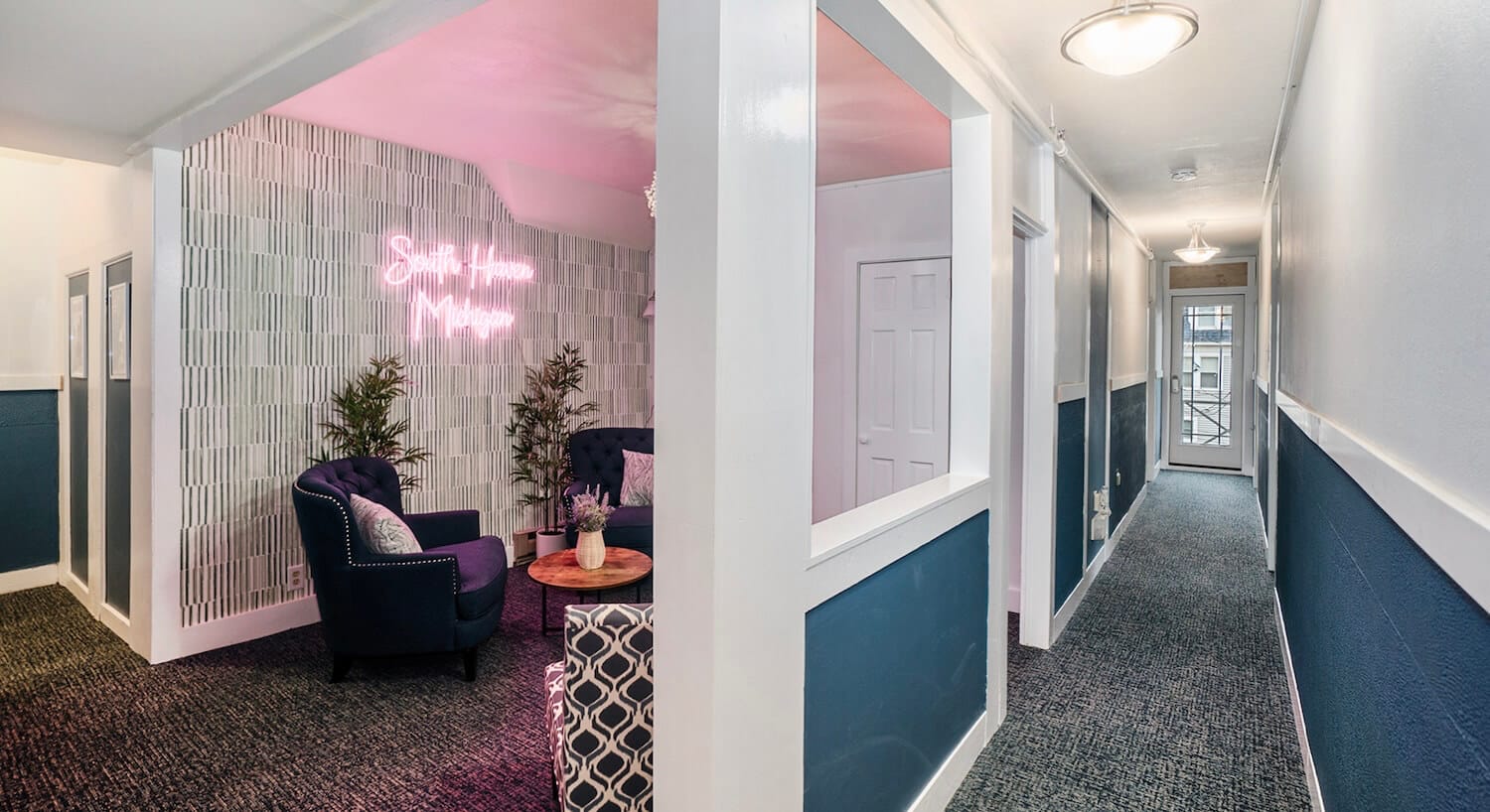 A long hallway with dark blue and white walls, and a sitting area off a doorway with a neon sign that says South Haven Michigan