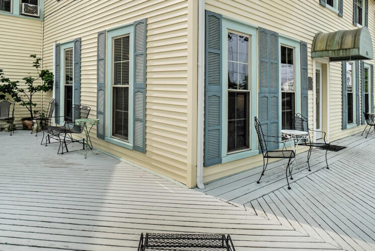 A pale yellow house with light green shutters next to the windows and deck areas with wrought iron tables and chairs.