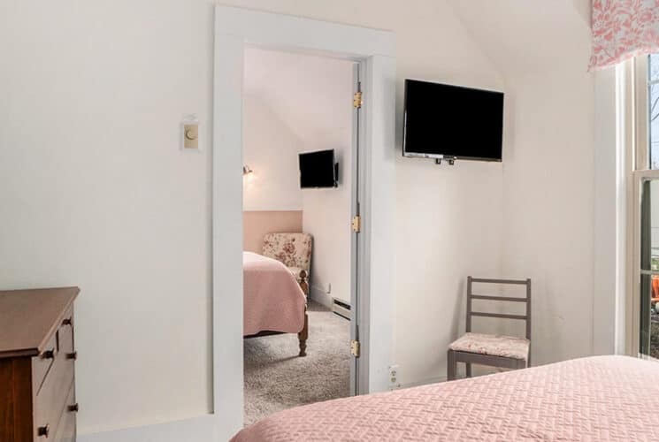Two adjoining bedrooms with beds with pink bedding, a wood dresser, a flat screen TV on the wall, and a window to outside.