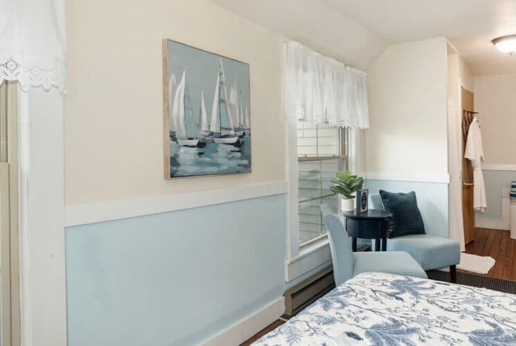 A bedroom with light blue and white walls, a painting of sailboats on the water on the wall, a corner sitting area with 2 plush chairs, and a corner tub in the background.
