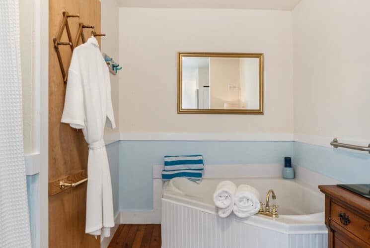 A bathroom with pale blue and white walls, a corner soaking tub, a bathrobe hanging from a hook on the back of a door, and a dresser along one wall.