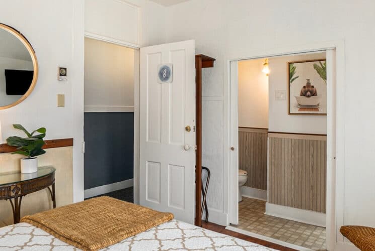 A bedroom with a half table along one wall with a lamp and mirror on the wall above it, open doorways to a private bathroom and the hallway, with #8 on the entry door.