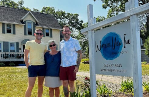 Two men and a women in front of a stately yellow house with blue shutters in front of a sign that says Waters View Inn Beach House.