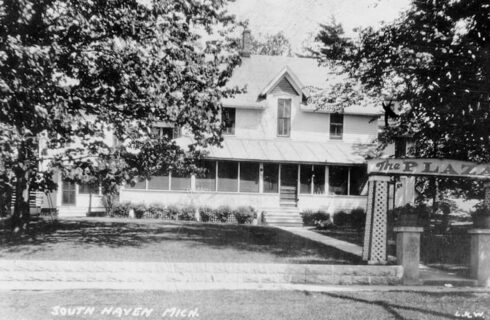 A black and whit photo of a stately inn with a screened porch spanning the width of the building, and a sign at the front of the sidewalk that says 