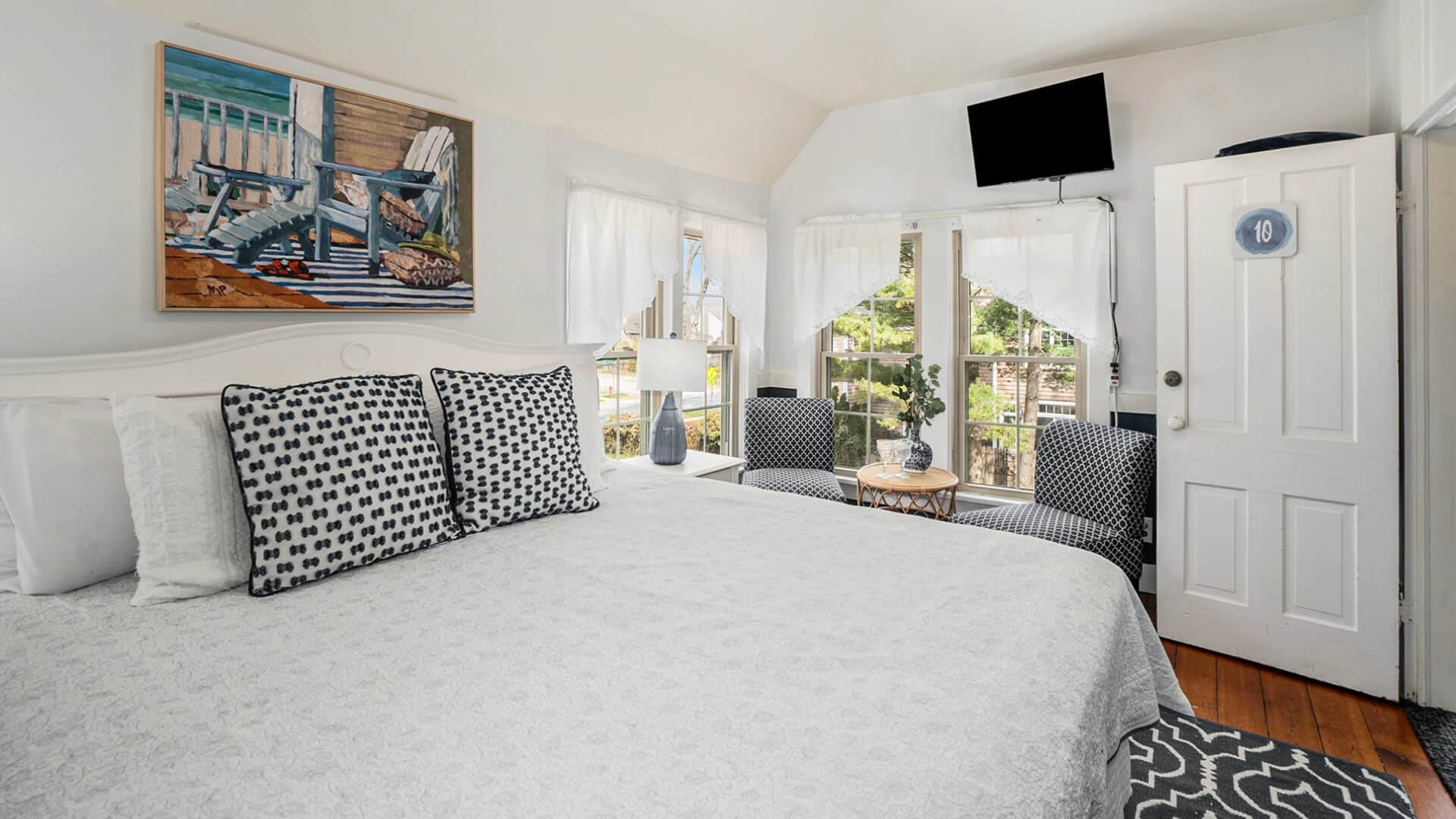 A bedroom with a bed with white bedding, blue accent pillows, wood floors and a sitting area in the corner along bright windows with 2 plush chairs and a small table between them, along with a flat screen TV on the wall above the windows. The #10 on the door.