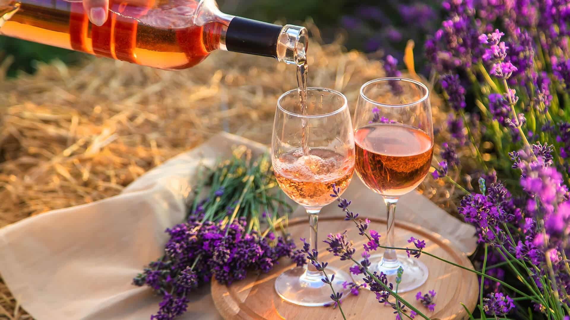 A bottle of blush pink wine being poured into 2 wine glasses on a wood tray with a handful of fresh purple flowers next to the tray.