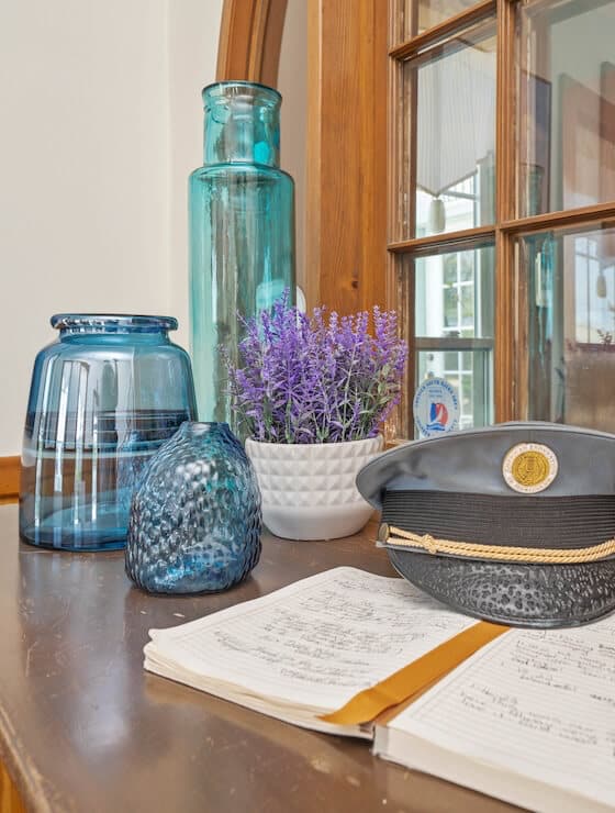 A wood table with a guest book, a sea captain's hat, some blue vases, and a white pot of purple flowers.