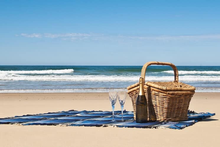 A wicker picnic basket, a bottle of champagne, and 2 champagne flutes on a blanket on the beach in front of the ocean.