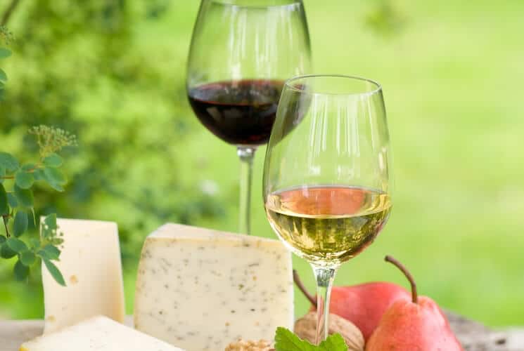 Two glasses of wine, one full of red wine and one with white wine, along with wedges of cheese and fresh fruit.